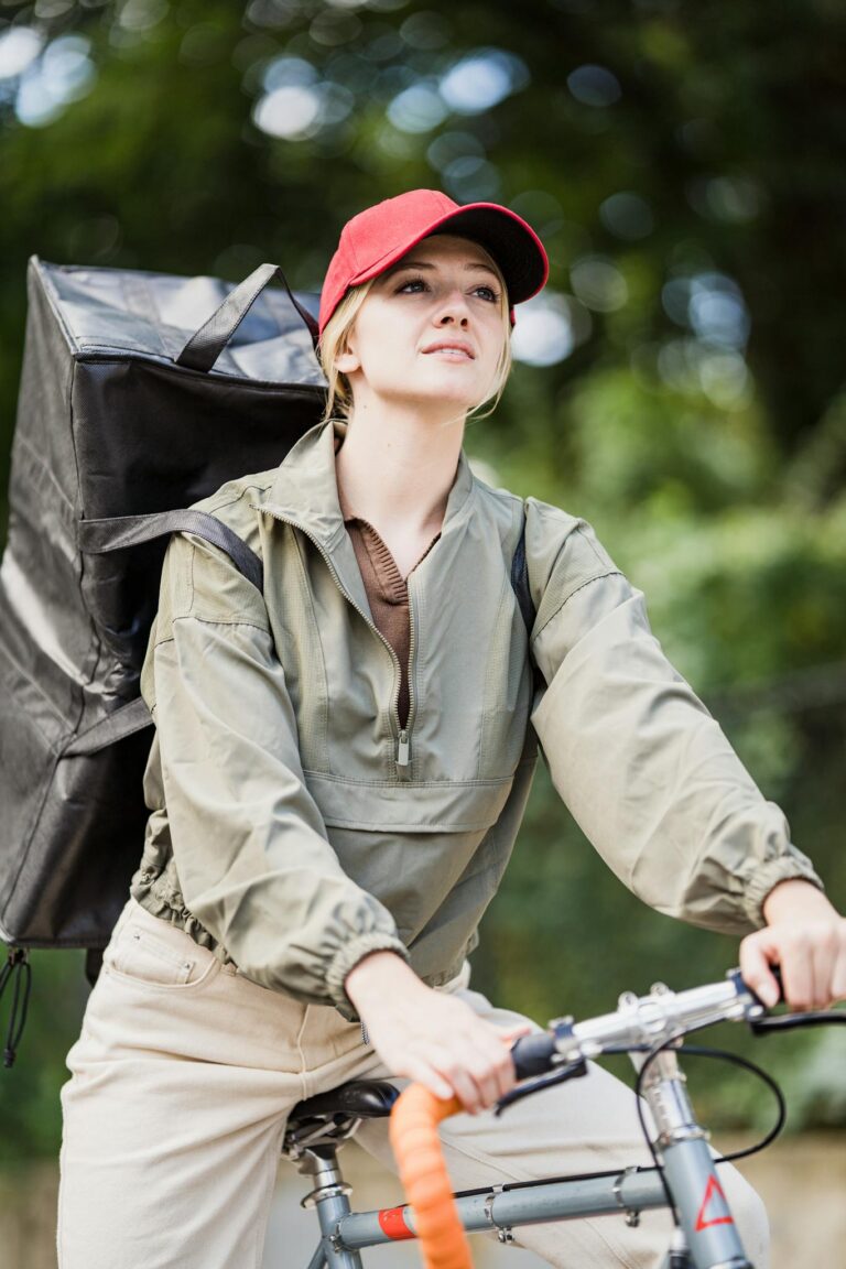 A female courier riding a bicycle with a large black delivery backpack outdoors.
